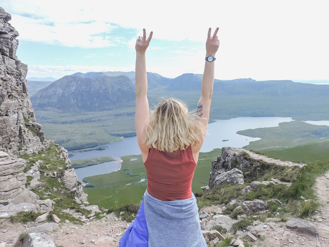 Gemma back to camera with arms in air with Stac Pollaidh mountain backdrop Scotland