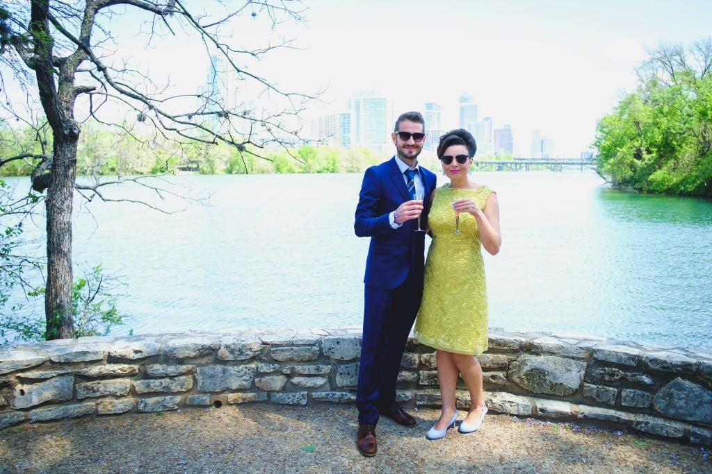 Bride in yellow dress, groom in navy suit, holding champagne flutes against Lady Bird Lake Austin backdrop