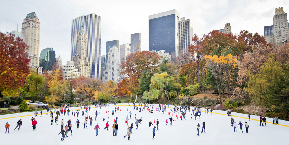 Ice skaters at Wollman Rink in Central Park NYC with fall trees and building in background